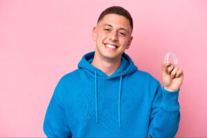 young Brazilian man holding invisible braces isolated on pink background smiling a lot